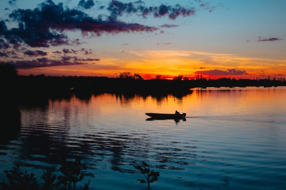 Maisemakuva järvenrannasta, jossa on tumma siluetti pienestä kalastajan moottoriveneestä vedessä juuri ennen auringonlaskua