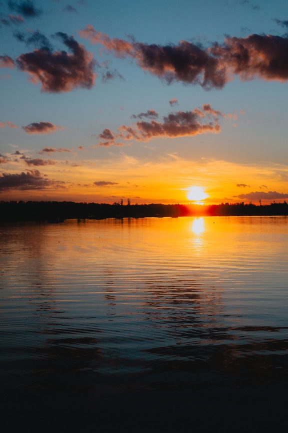 Sonnenuntergang über einem Seeufer mit orange-gelbem Sonnenlicht, das sich auf ruhigem Wasser spiegelt