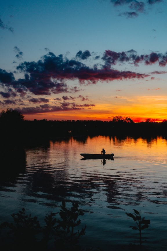 Ihmisen siluetti pienessä moottoriveneessä järvellä auringonlaskun aikaan, tumman oranssinkeltaisella auringonvalolla, joka heijastaa järven rauhallista vettä