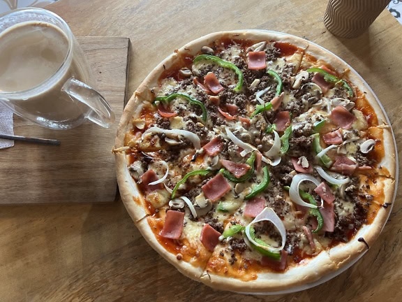 A gourmet pizza with meat and vegetables on a wooden table next to coffee mug in pizzeria