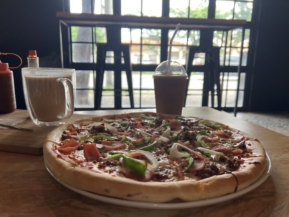 Gourmet pizza with vegetables and meat toppings on a plate next to a coffee mug on table in pizzeria