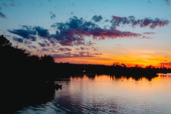 Auringonnousu järven rannalla heijastaen nousevaa aurinkoa tyynellä vedenpinnalla