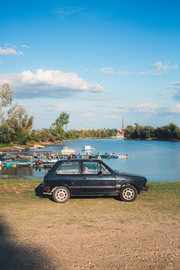 Vista lateral de um carro Zastava Yugo 45, automóvel estacionado ao lado de um lago com marina para pequenas lanchas ao fundo