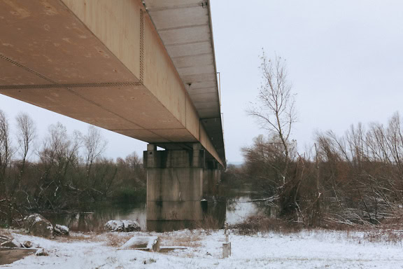 Underneath of an old concrete bridge over water with snow on the ground