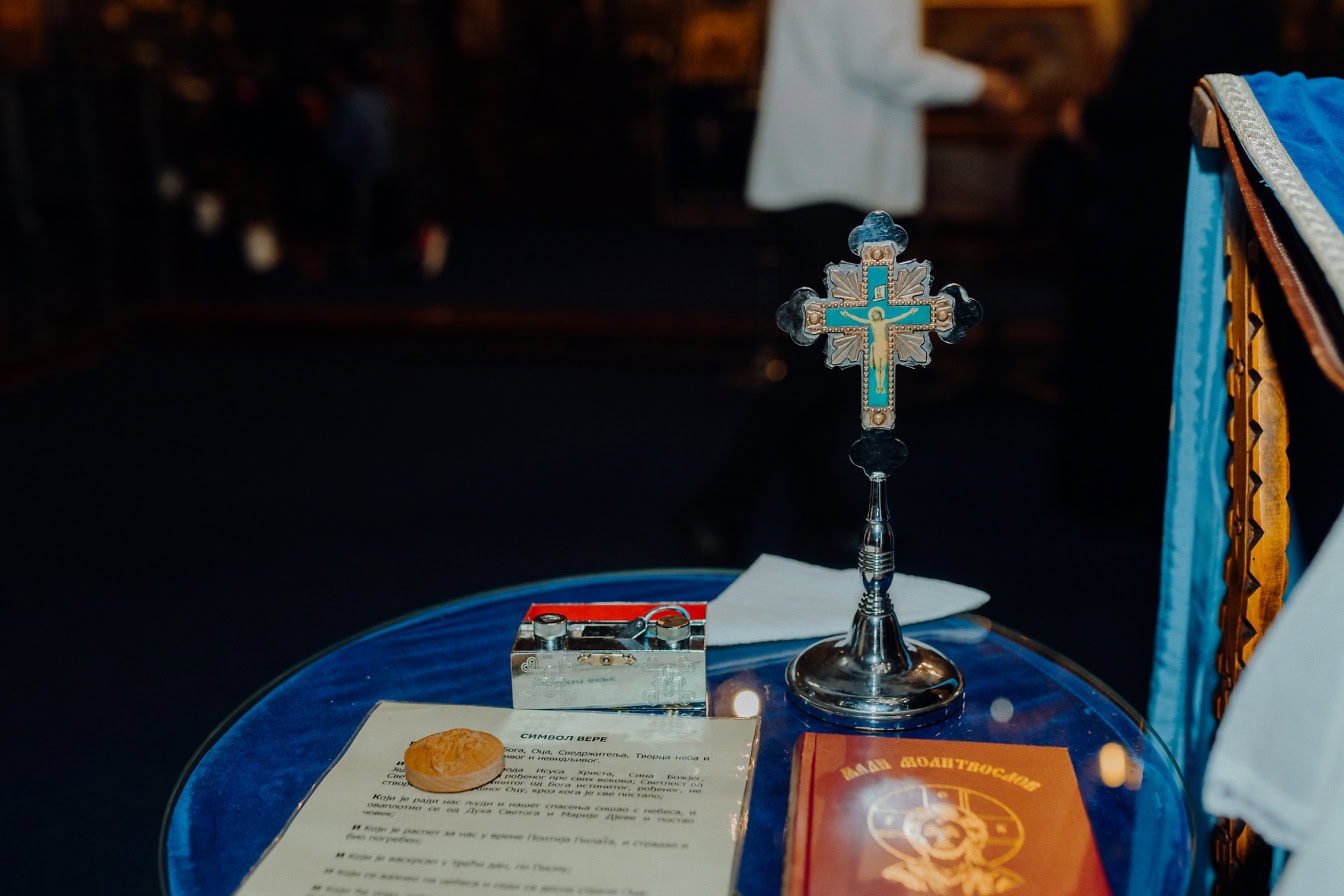 An ornate metal Christian cross depicting the crucifixion of Jesus Christ on a table next to a holy book and other sacred religious objects