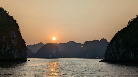 베트남의 하롱 베이 (Ha Long bay) 위로 흐릿한 태양과 함께 장엄한 일몰과 함께 섬과 산의 실루엣이 있습니다.