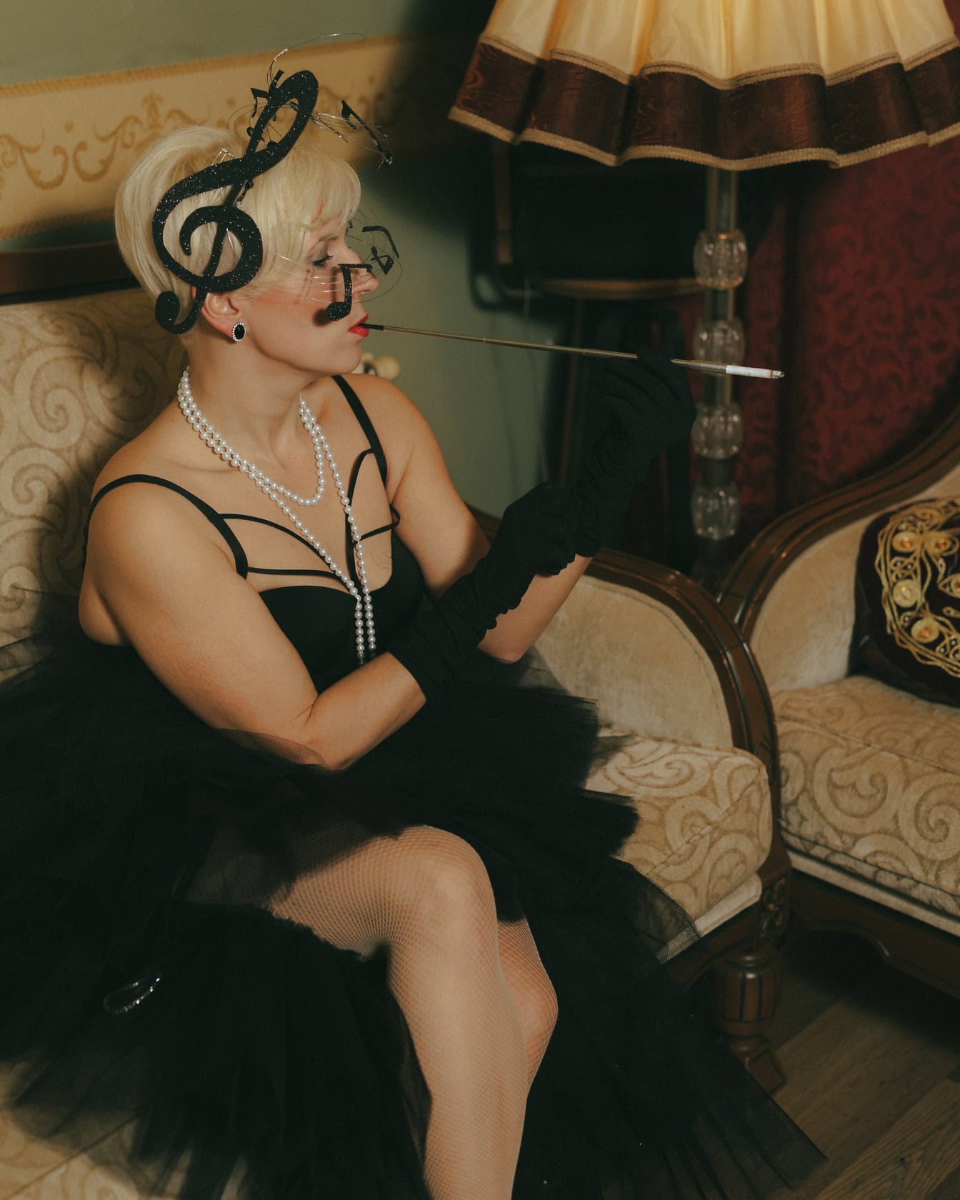 A lady sitting on a couch in elegant black dress, gloves and hat and smoking cigarettes with fancy cigarette holder