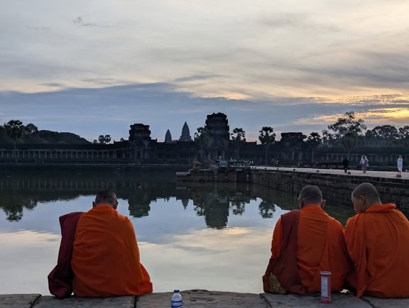 Buddhista szerzetesek hagyományos köntösben ülnek egy kőtömbön a víz mellett, háttérben az ősi Angkor Vat templommal, Kambodzsa