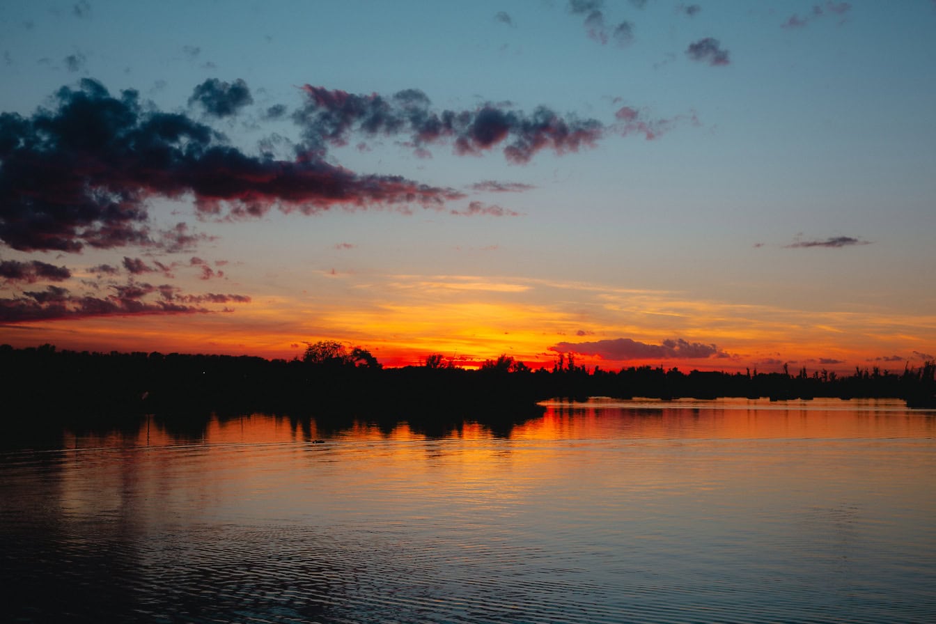 Fotografia di paesaggio scattata poco prima dell’alba sul lago con un drammatico cielo rosso-arancio e nuvole scure, un’immagine di paesaggio vivida e satura