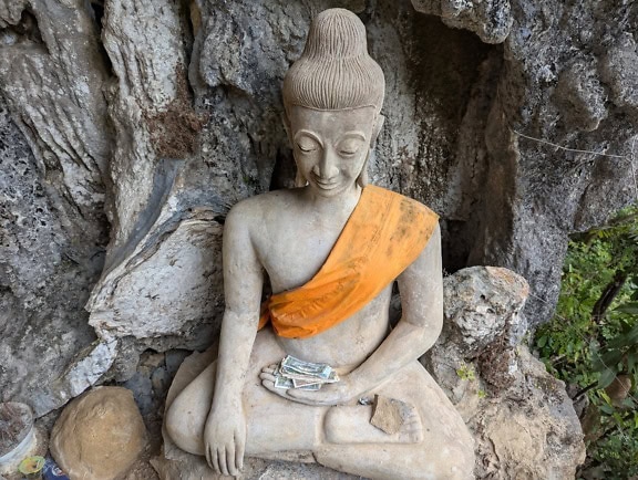 Stone statue of Buddha sitting and meditating in lotus position with monetary offerings, Buddhist sculpture near Vang Vieng town, Laos